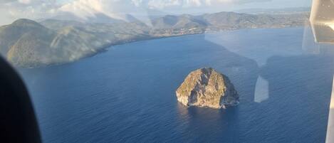 An aerial view of a coastal landscape with a prominent rocky island in the center, surrounded by calm blue waters and distant mountains.