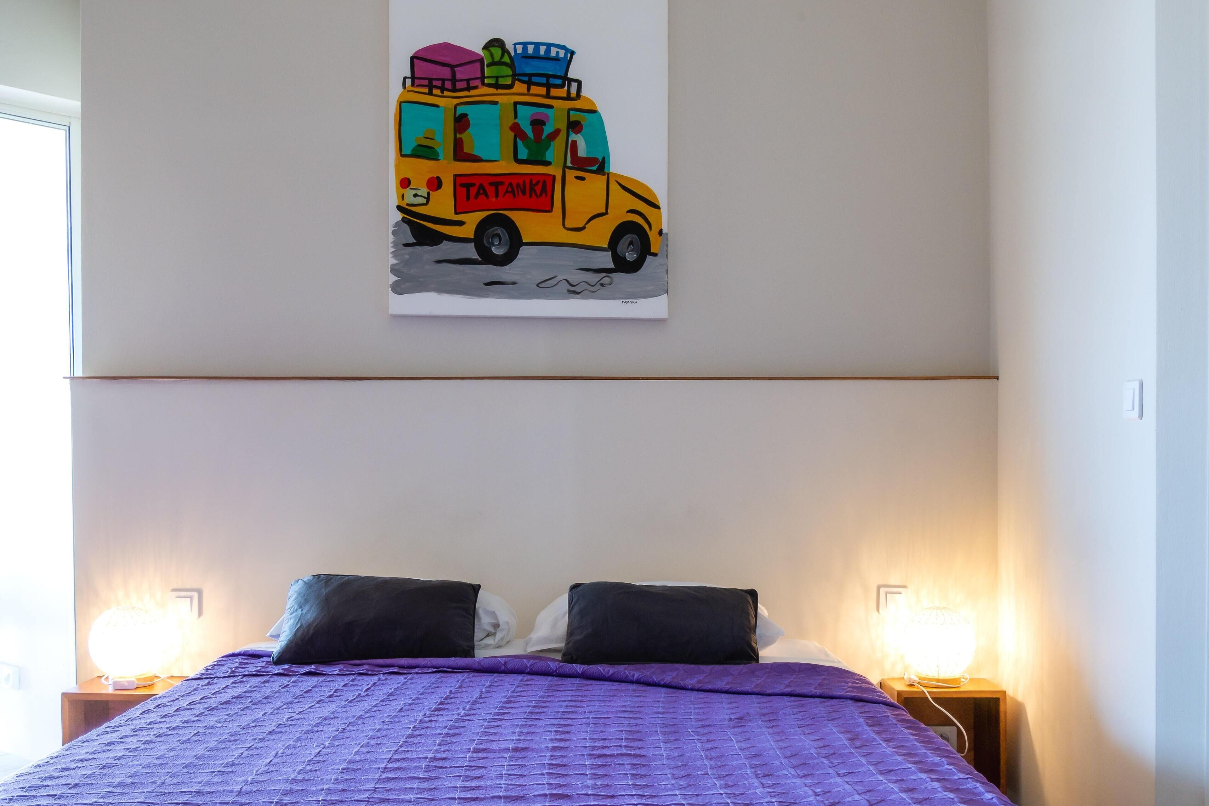 A bed with a purple quilt, two black pillows, and a wall-mounted painting of a yellow van with passengers and luggage.