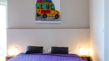 A bed with a purple quilt, two black pillows, and a wall-mounted painting of a yellow van with passengers and luggage.