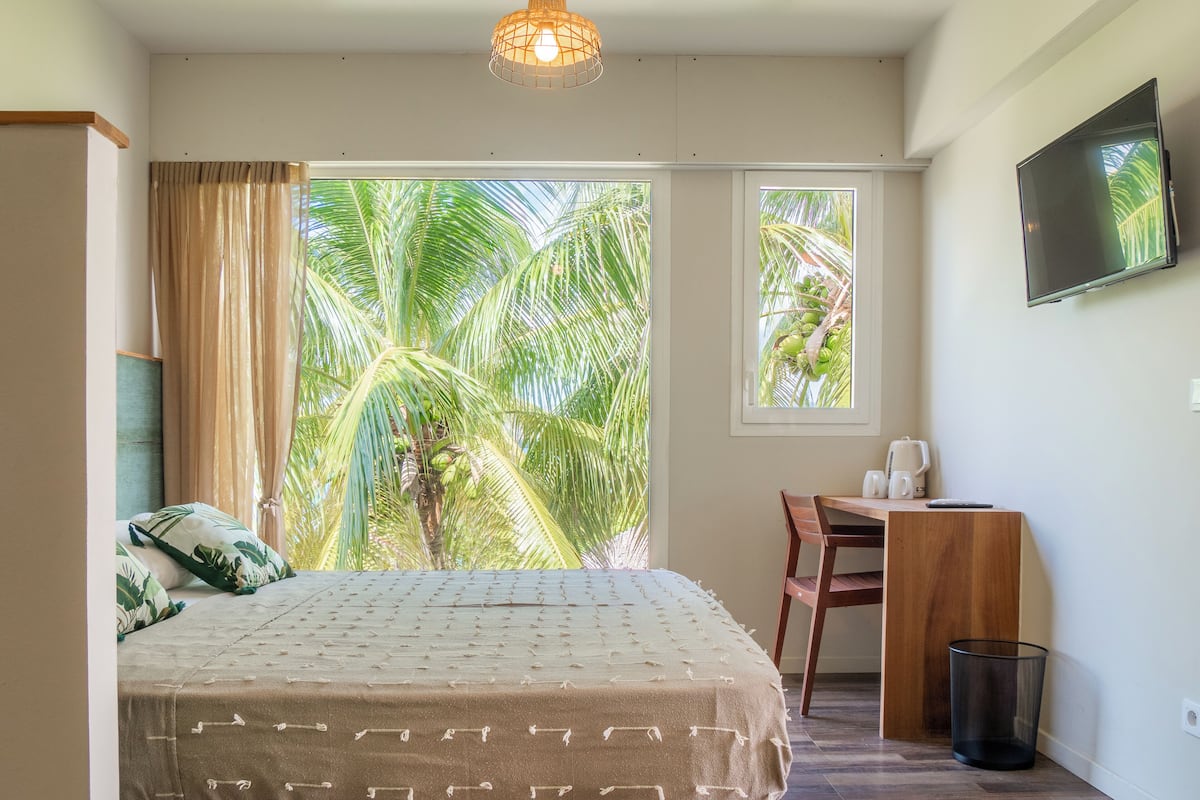 A bedroom with a bed, a desk, a chair, a TV, and a window with a view of palm trees.