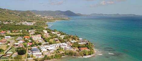 An aerial view of a coastal town with houses along the shoreline, a clear blue sea, and distant mountains.
