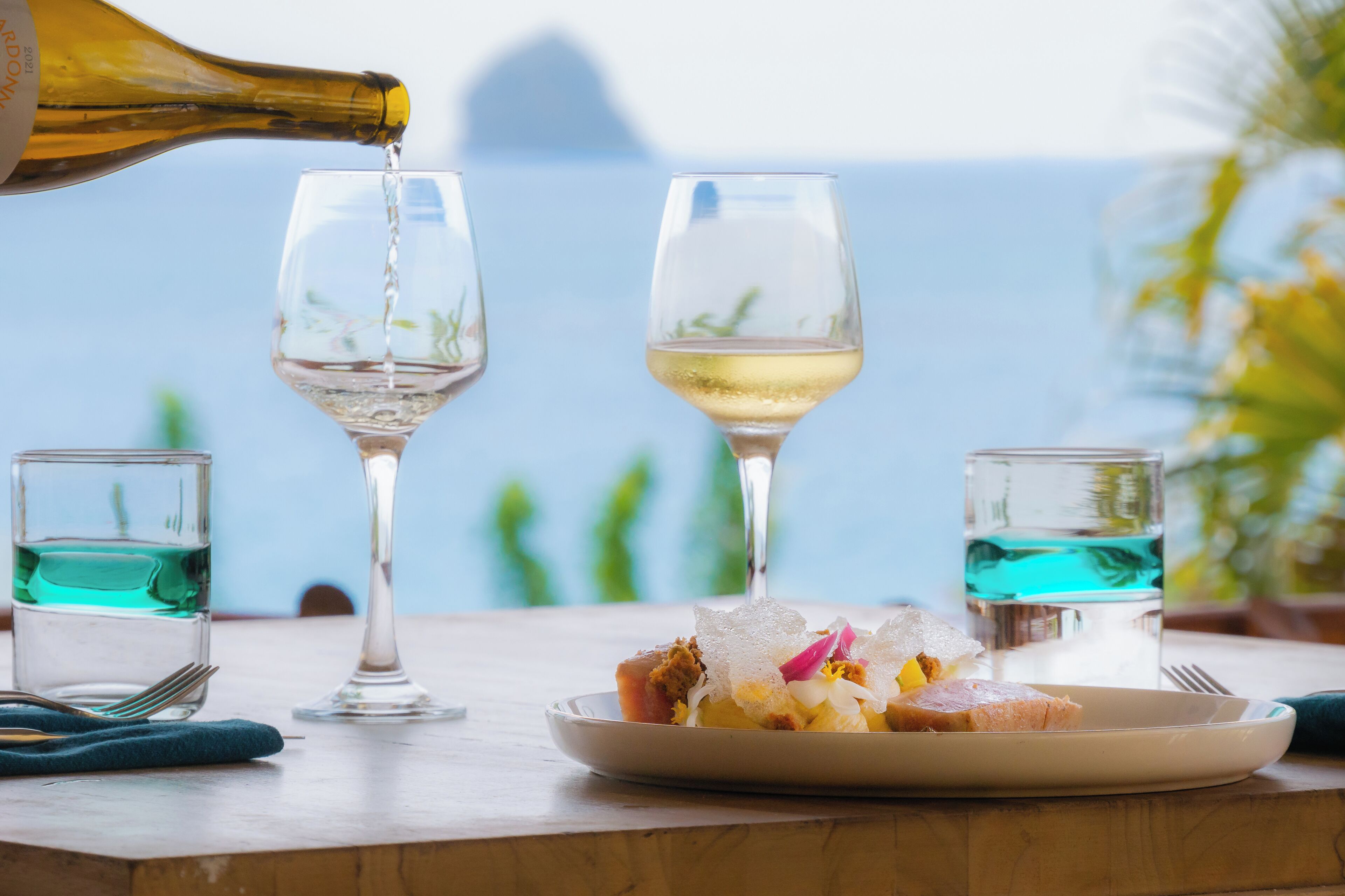 Una mesa con un plato de comida, dos copas de vino y una botella vertiendo vino.