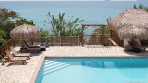 Un área de piscina con sillones de descanso y sombrillas de paja, con vista a un mar tranquilo y islas distantes.