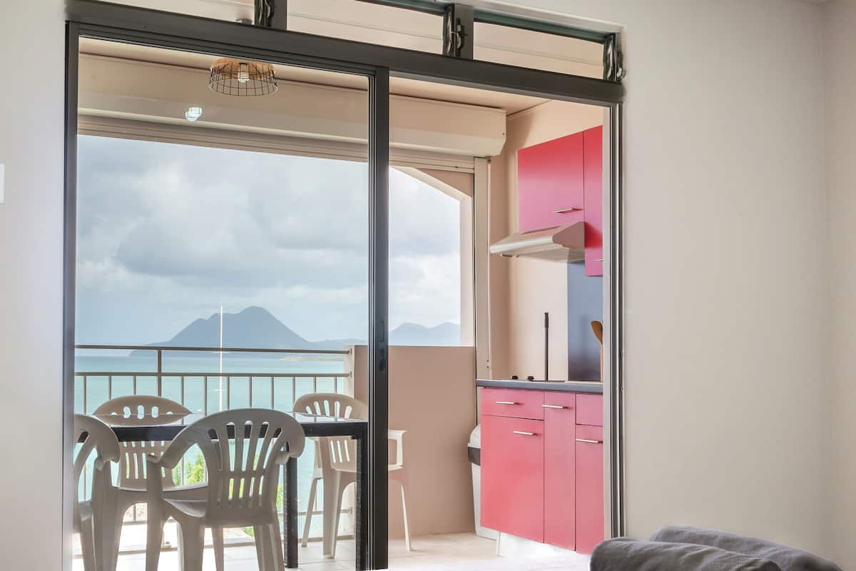 A bedroom with a bed, a balcony with chairs, and a view of the sea.