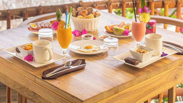 Una mesa de comedor de madera dispuesta con alimentos para el desayuno, incluyendo un tazón de fruta, un vaso de jugo de naranja y un plato de comida, con vista a una playa y una estructura de techo de paja.