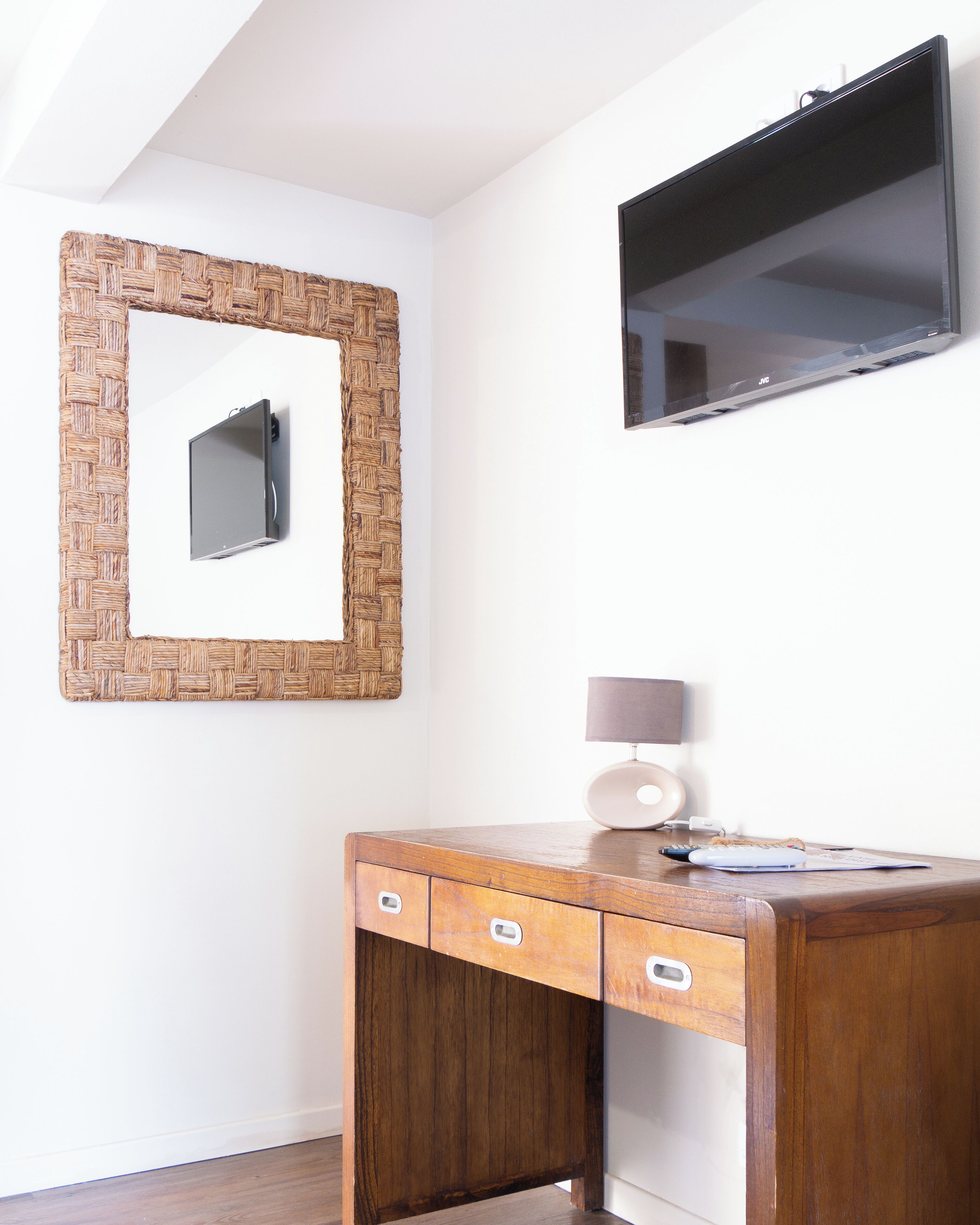 A wooden desk with drawers and a lamp, a woven mirror, and a flat-screen TV mounted on the wall.