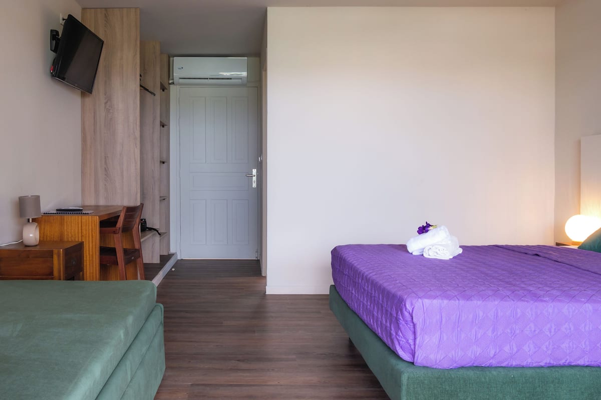A hotel room with a bed, a desk, a chair, a TV mounted on the wall, and an air conditioning unit.