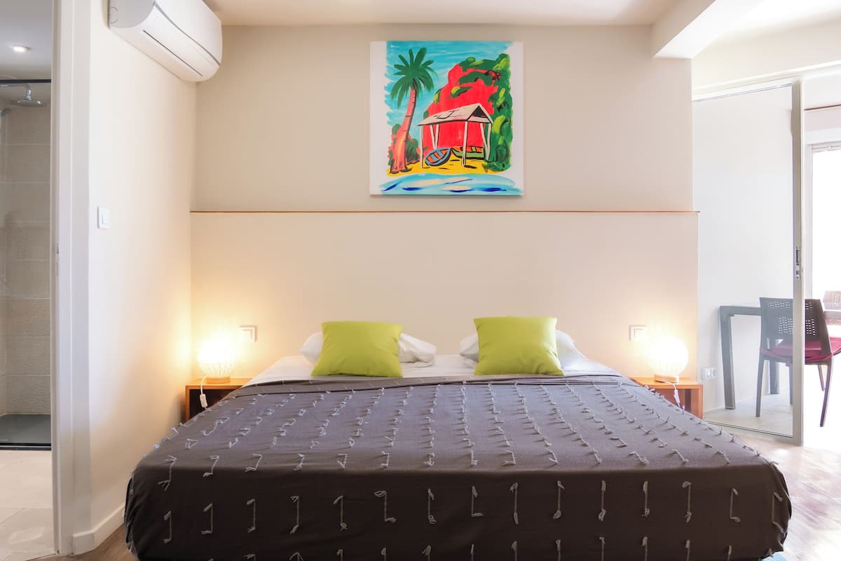 A hotel room with a bed, two green pillows, a painting of a beach scene, and a chair in the corner.