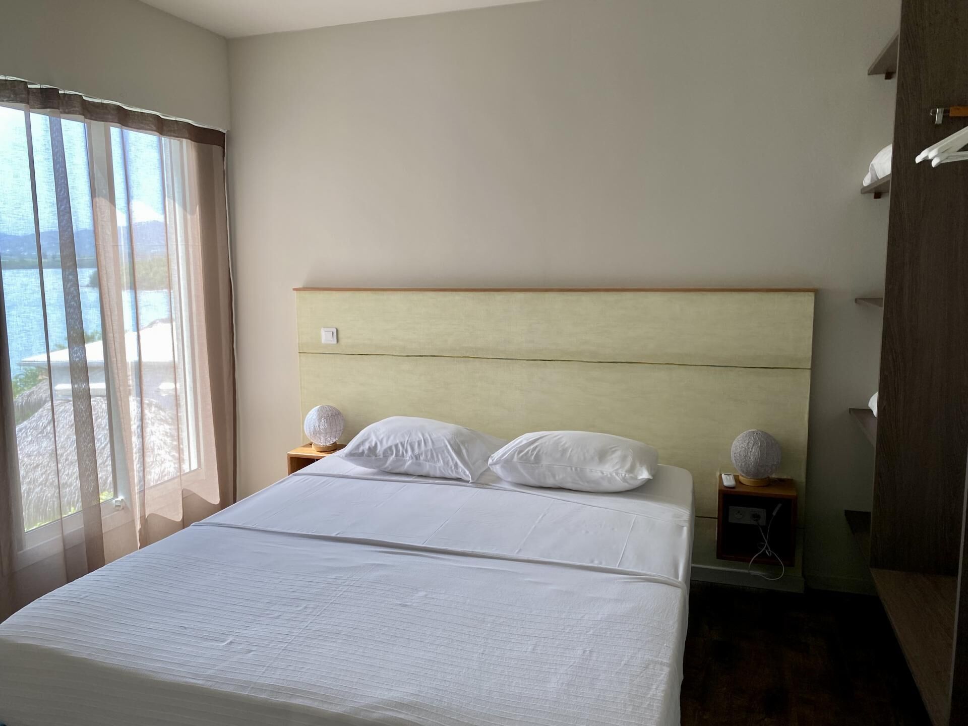 Une chambre d’hôtel avec un grand lit, deux tables de chevet et une vue sur la mer, visible à travers de fines rideaux.