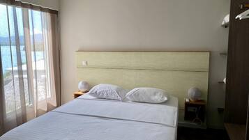 Une chambre d’hôtel avec un grand lit, deux tables de chevet et une vue sur la mer, visible à travers de fines rideaux.