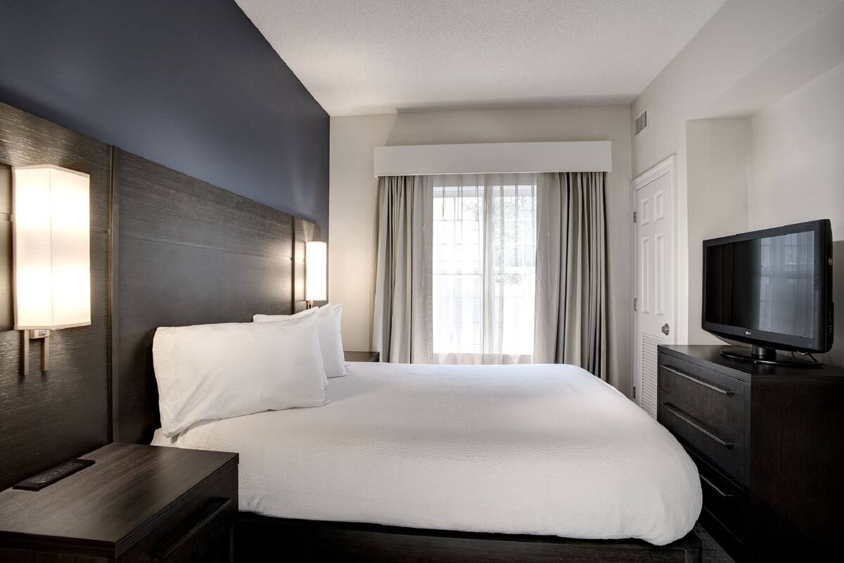 Habitación de hotel con una cama grande, televisión de pantalla plana, mesita de noche y ventana con cortinas.