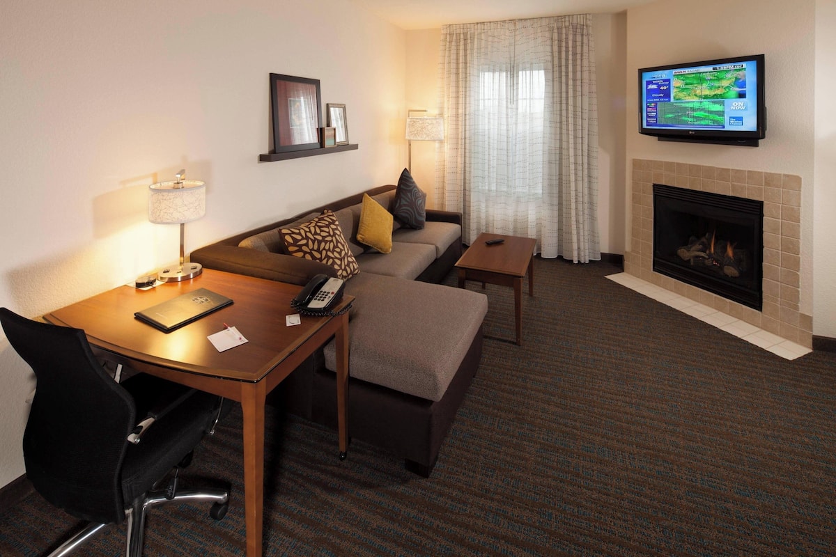 A hotel room with a sofa, a small table with a lamp, a TV mounted above a fireplace, and a carpeted floor.