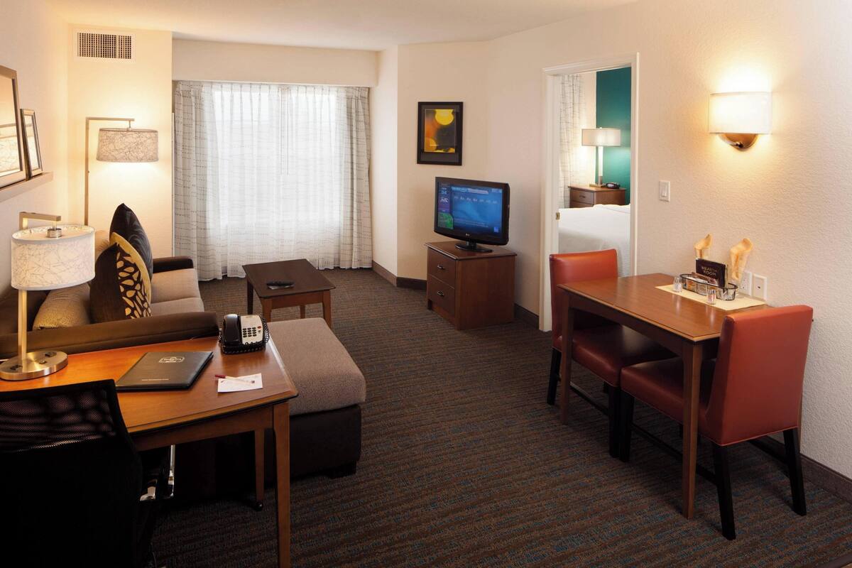 A hotel room with a sofa, a desk with a laptop, a television, a dining table with chairs, and a window with curtains.