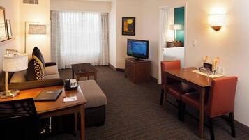 A hotel room with a sofa, a desk with a laptop, a television, a dining table with chairs, and a window with curtains.