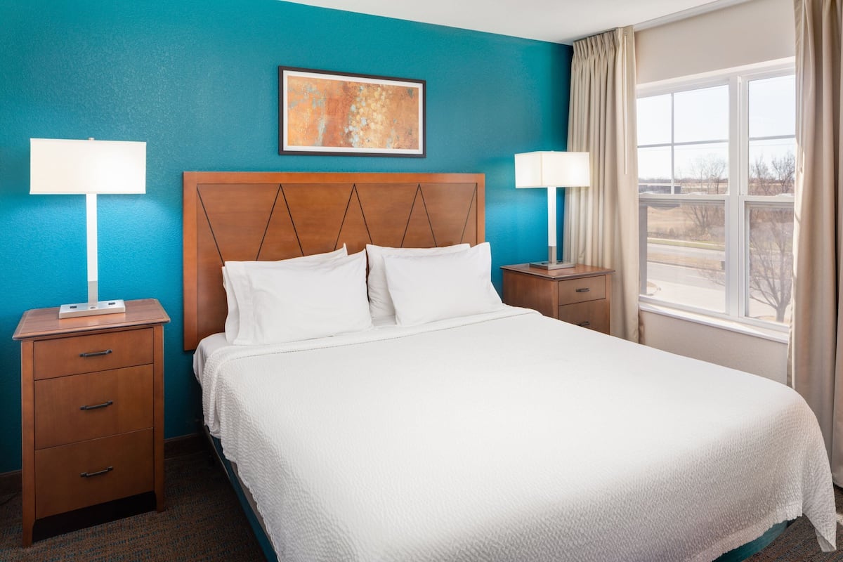 A hotel room with a large bed, two bedside tables, a nightstand, a window with curtains, and a painting on the wall.