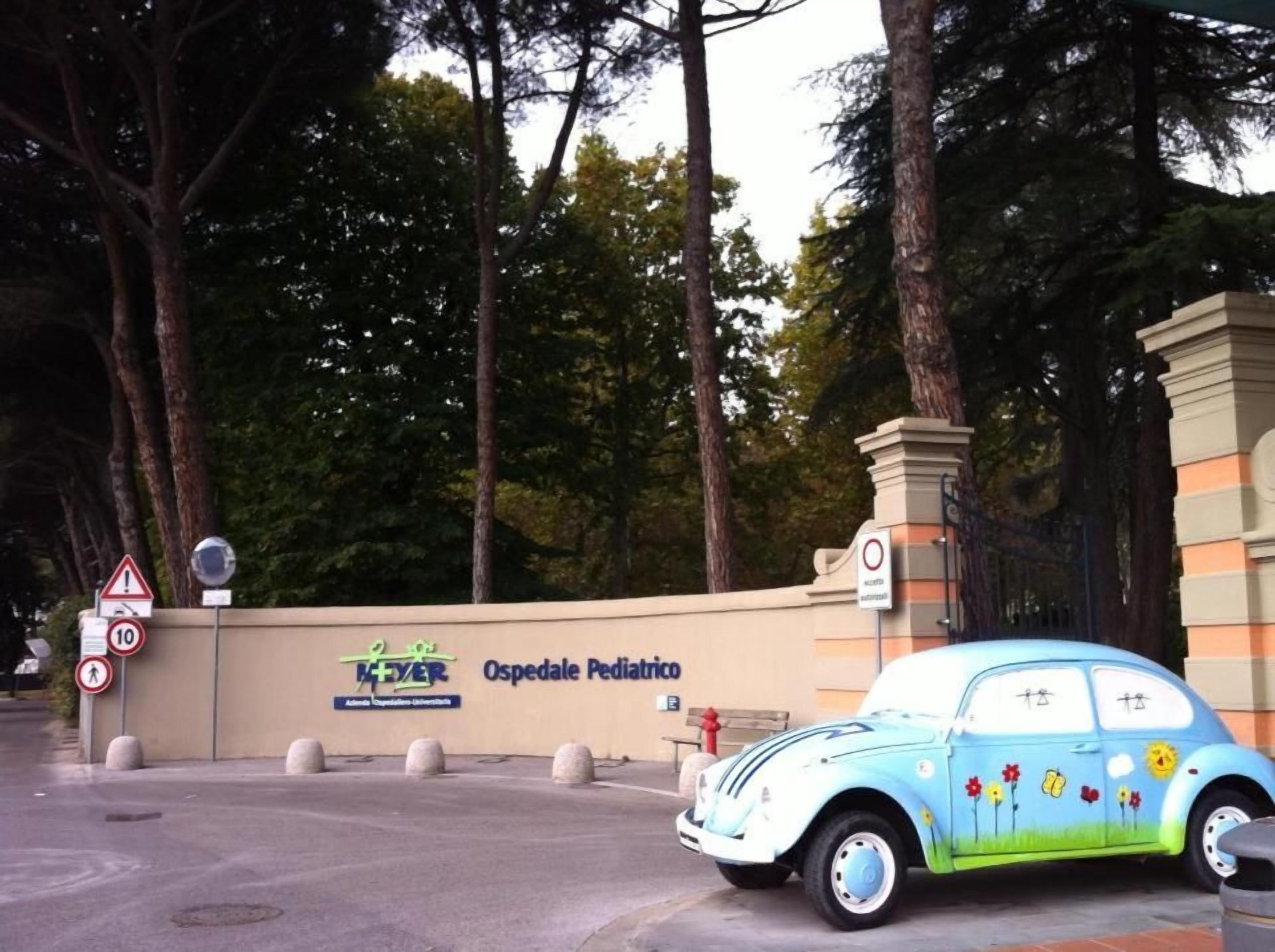 A pediatric hospital entrance with a blue car parked in front, a sign for Ospedale Pediatrico, and traffic signs including a speed limit of 10.
