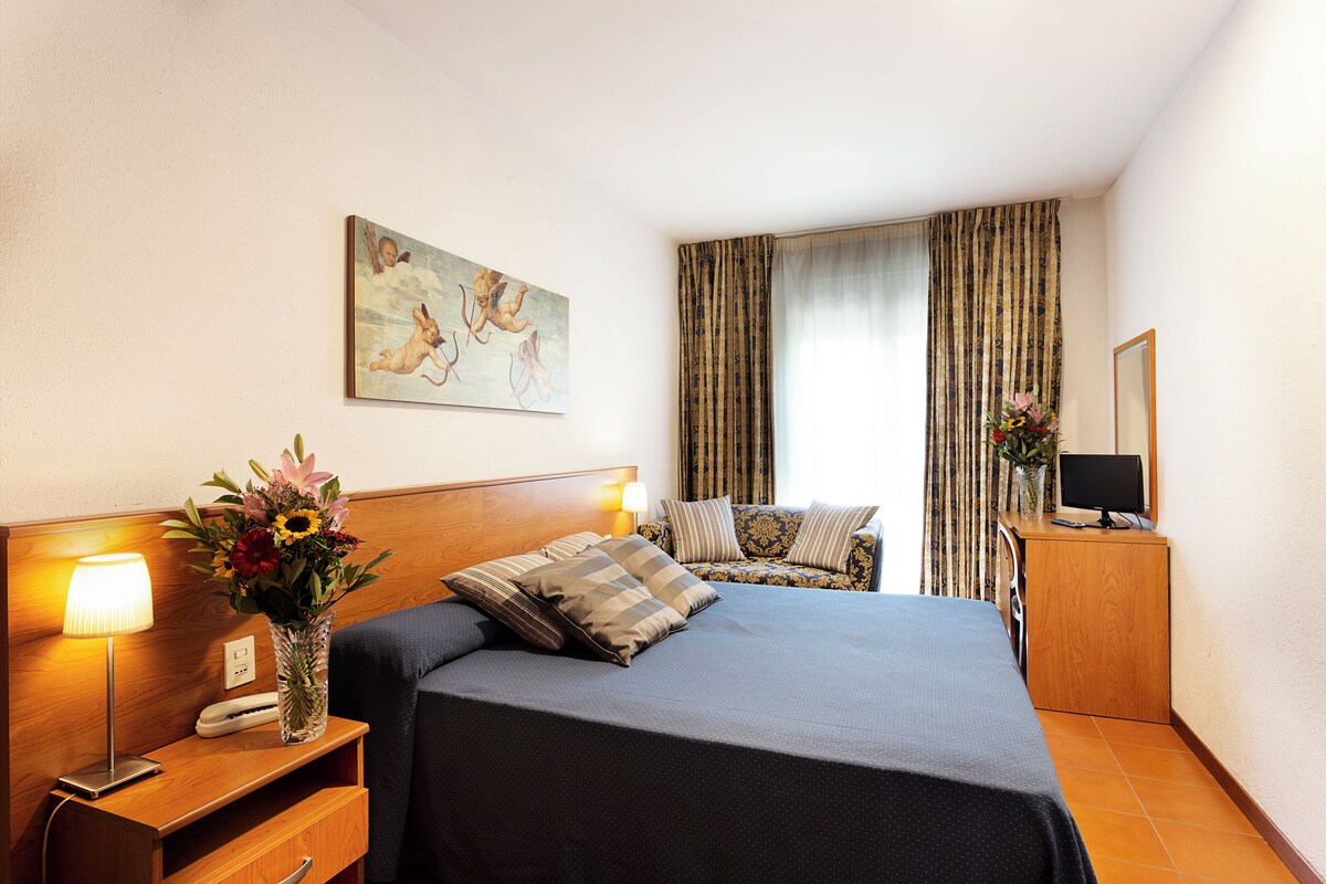 Habitación de hotel con cama, lámparas de noche, un escritorio con computadora, un florero con flores y un cuadro en la pared.
