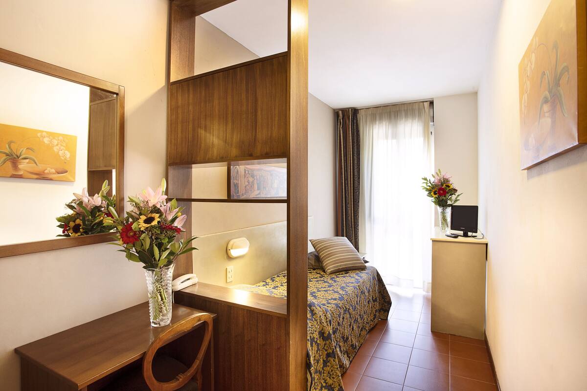 Habitación de hotel con escritorio de madera, una silla, cama con colcha estampada, mesita de noche con lámpara y una pequeña planta en maceta.