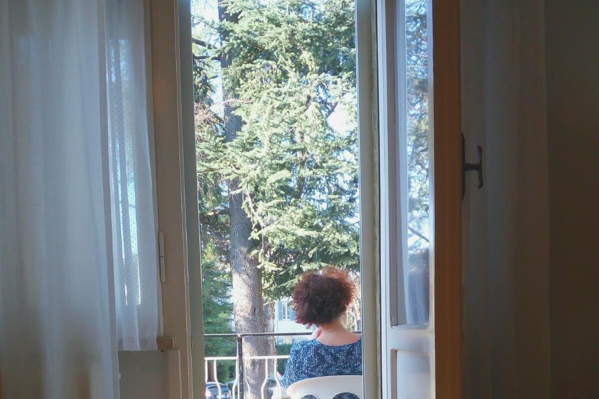 A room with a bed, a window with curtains, and a view of trees and a fence outside.