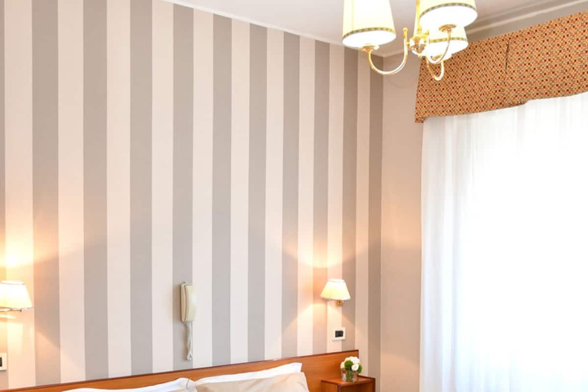A hotel room with a large bed, striped wallpaper, a chandelier, and a bedside table with a lamp.