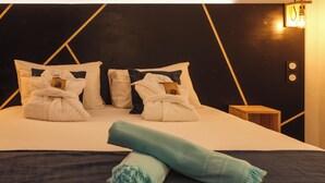Una cama bien tendida con almohadas y una toalla enrollada, una mesita de noche con lámpara y una pared con motivos geométricos.