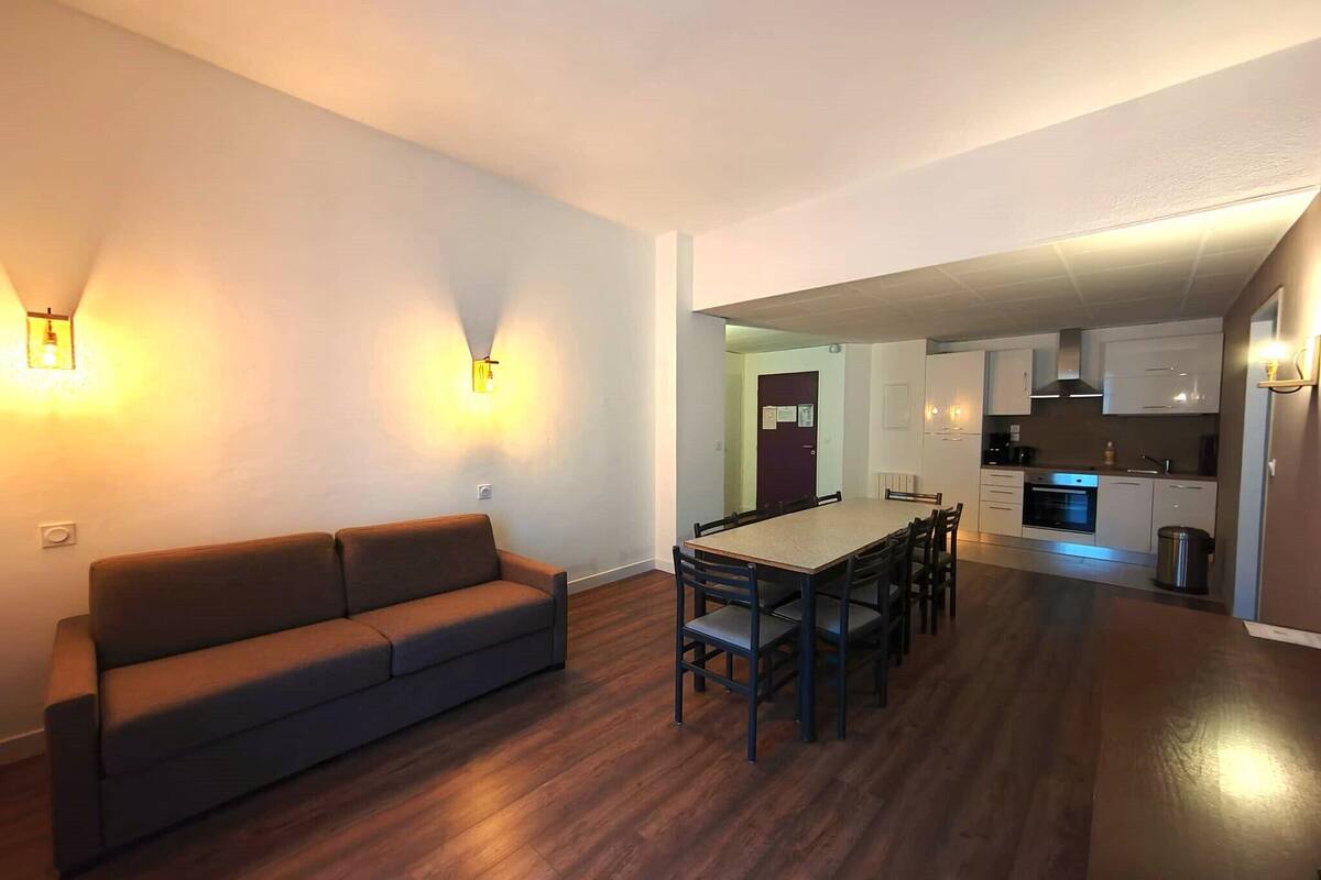 A modern living room with a sofa and dining table, a kitchen area with cabinets and appliances, and a hallway leading to another room.