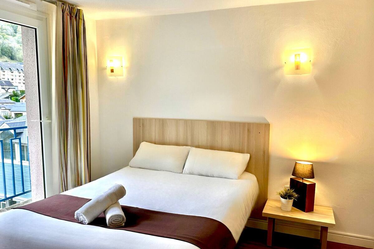 A hotel room with a bed, a nightstand with a lamp, and a view of buildings outside.