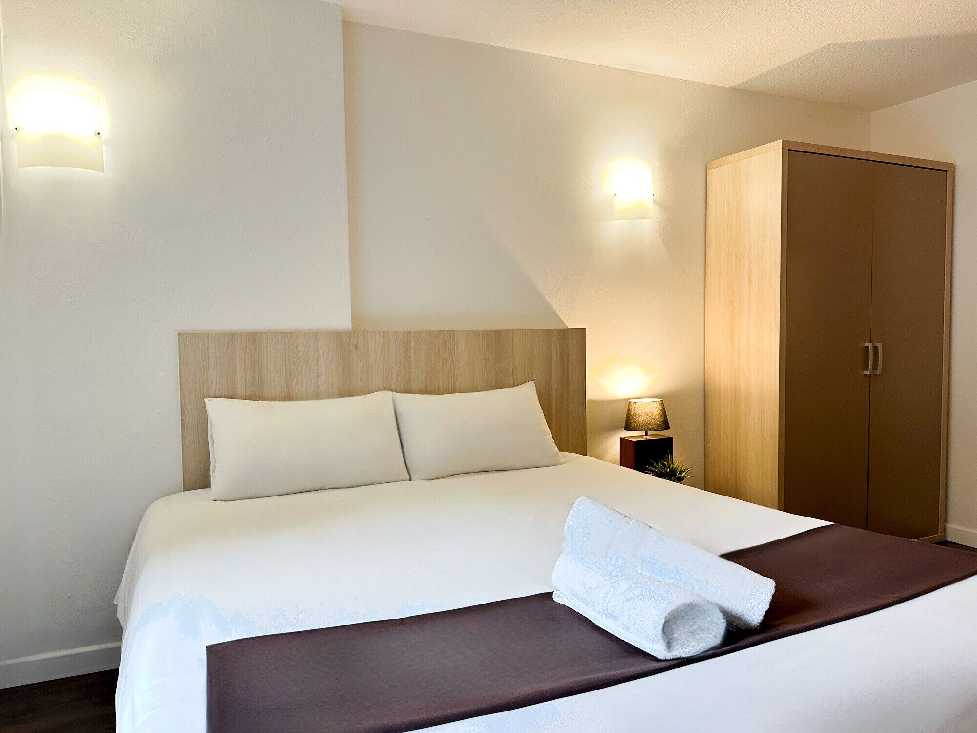 Un letto rifatto con una coperta marrone, un cuscino bianco e un asciugamano arrotolato sopra. A destra si trova un'armadio in legno e, alla sinistra, un comodino con una lampada.