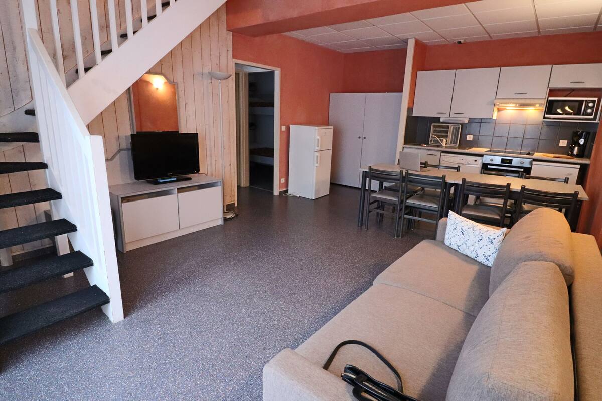 A modern kitchen with white cabinets, a microwave, and a stove. There is a dining area with a table and chairs, and a living room with a beige sofa and a TV on a stand.