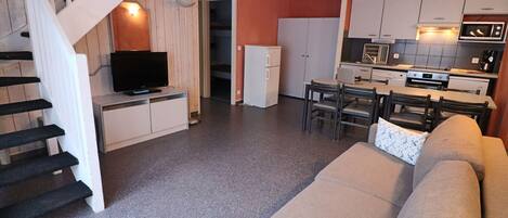 A modern kitchen with white cabinets, a microwave, and a stove. There is a dining area with a table and chairs, and a living room with a beige sofa and a TV on a stand.