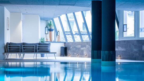 Una piscina interior con agua azul cristalina, diseño arquitectónico moderno y zona de estar con sofá y plantas en macetas.