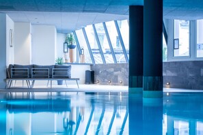 An indoor swimming pool with clear blue water, a modern architectural design, and a seating area with a sofa and potted plants. - Garden & City Cauterets Balneo (Cauterets)