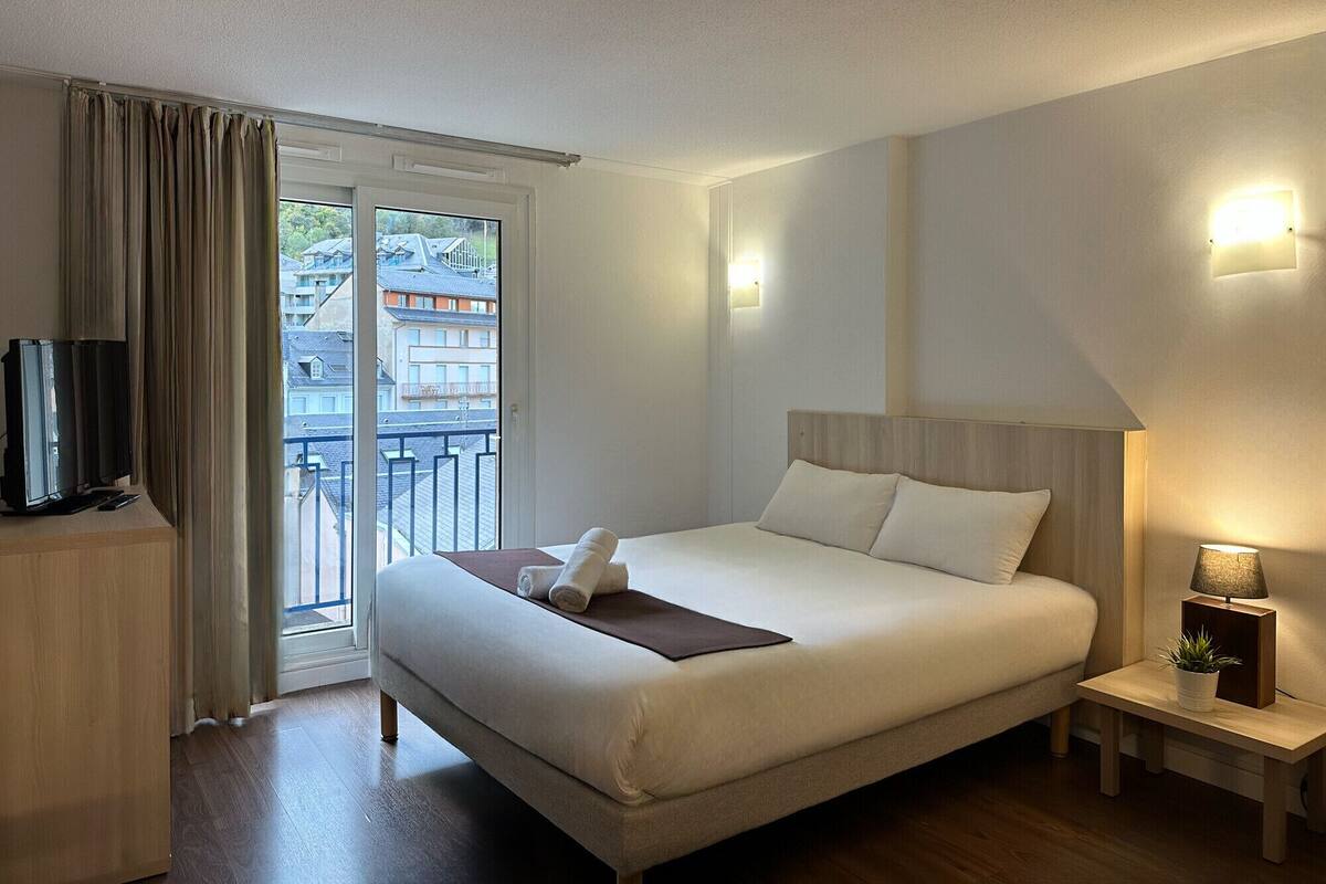 A hotel room with a bed, a television, a nightstand, and a view of the outside.