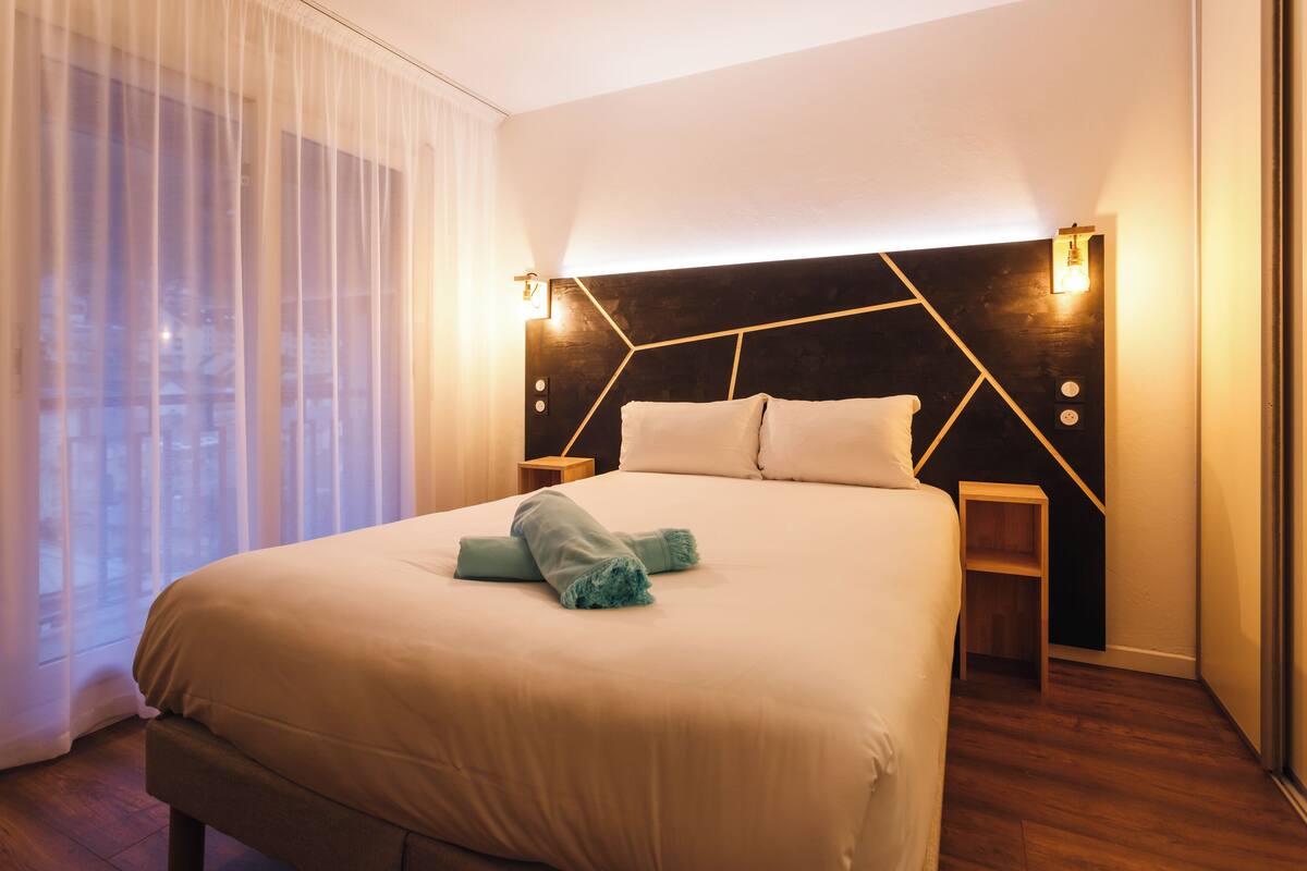 A neatly made bed with a geometric headboard, bedside tables, and a view of the city through sheer curtains.
