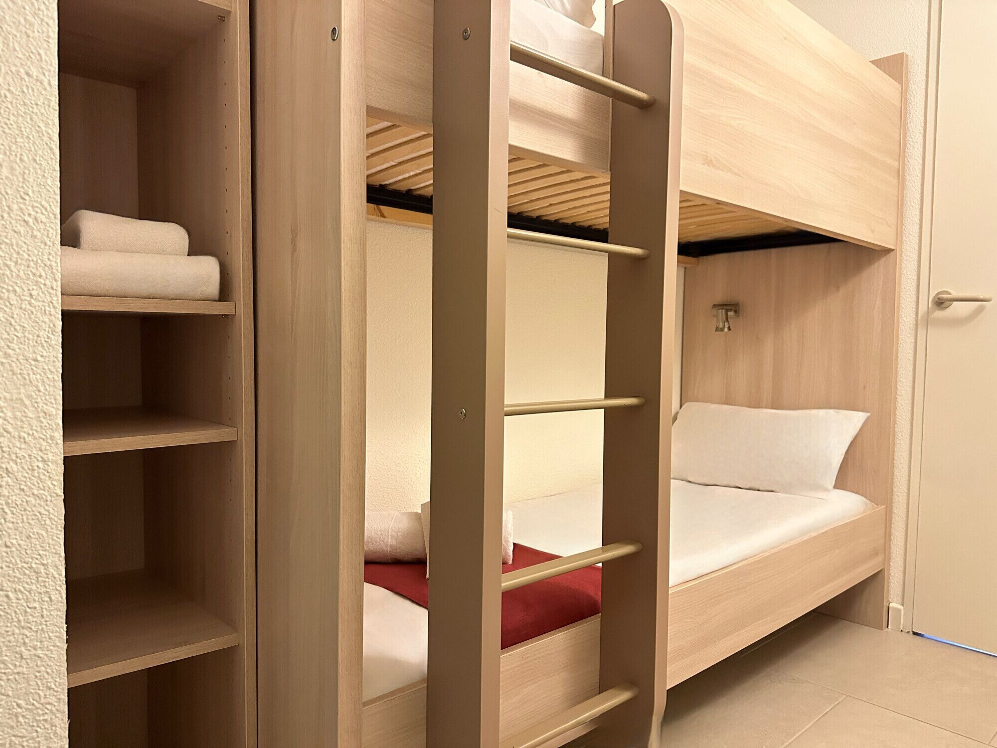 Una cama litera con escalera, un almohadón rojo y una manta blanca.