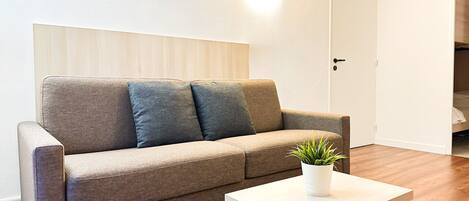 Un sofá de tonos neutros con dos cojines, una mesita blanca con una planta y suelo de madera.