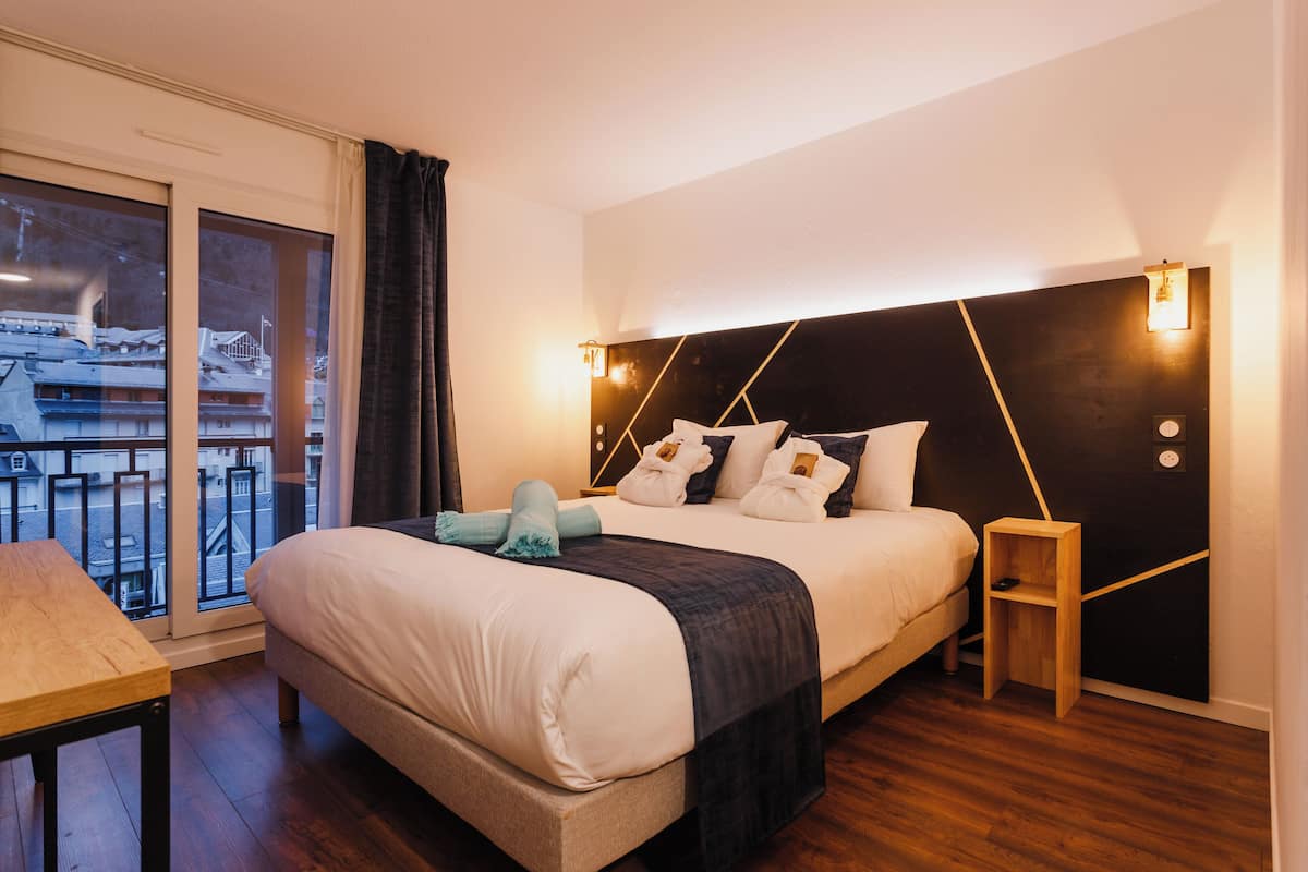 A modern hotel room with a large bed, a small bedside table, a view of the city through a large window, and a balcony.