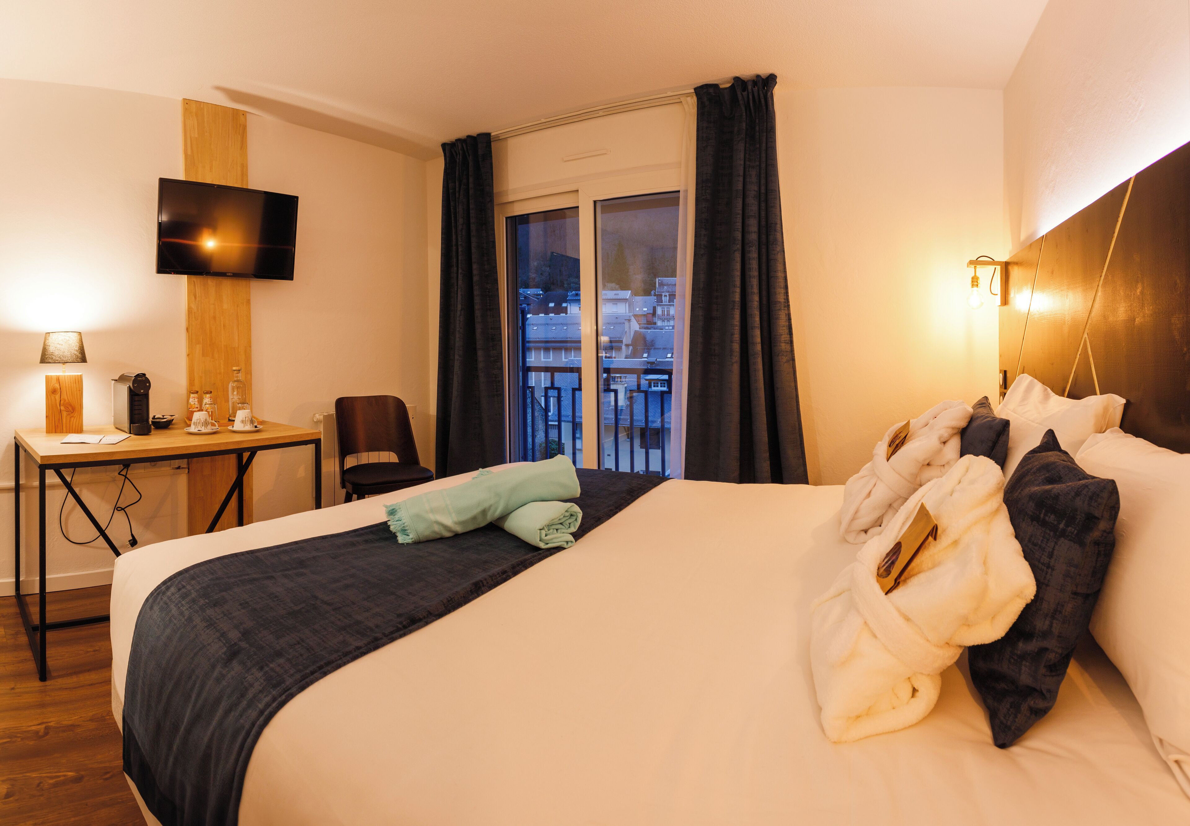 Quarto de hotel com uma cama grande, uma escrivaninha com cadeira, uma televisão na parede e uma varanda com cortinas.