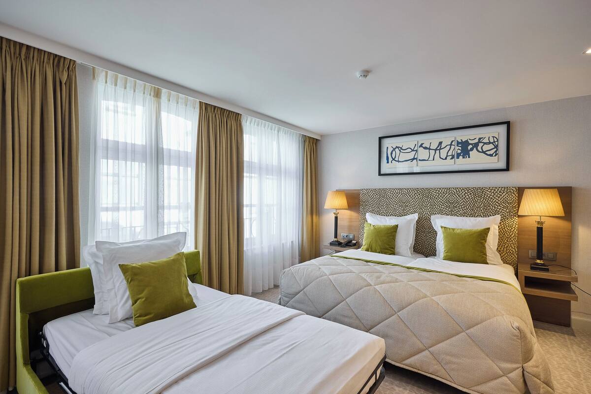 A hotel room with two beds, beige curtains, a framed artwork on the wall, and a nightstand with a lamp.
