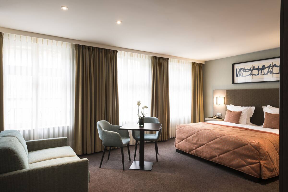 A hotel room with a bed, a sofa, a table with chairs, and a large window with curtains.
