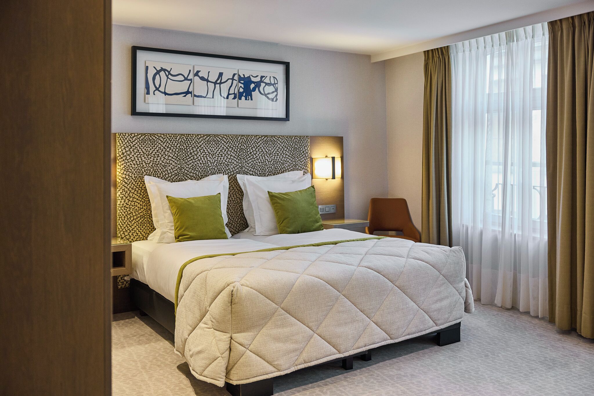 A hotel room with a large bed, a headboard with a patterned design, a bedside table with a lamp, and a window with curtains.