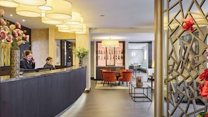 A hotel reception area with a dark reception desk, multiple light fixtures, and a seating area with orange chairs. - Aragon House (Bruges)