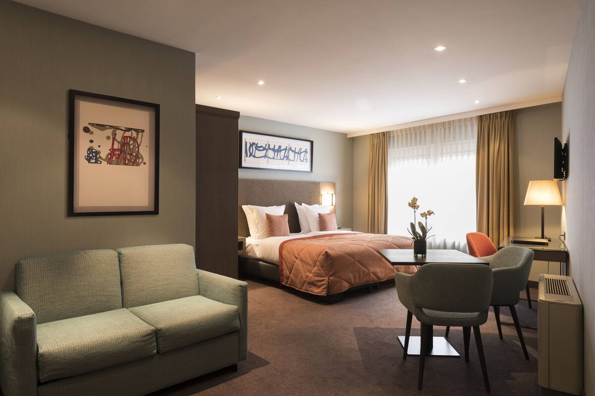 A hotel room with a bed, a sofa, a desk with a chair, a TV, and a framed picture on the wall.