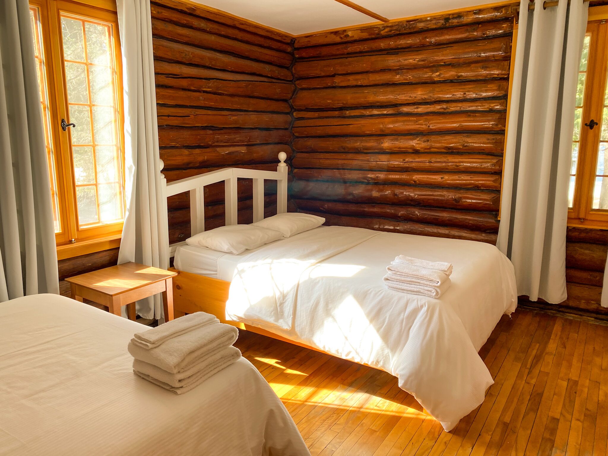 A log cabin-style bedroom with a single bed, a small bedside table, and a window with curtains.