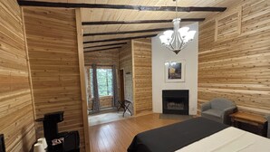 Interior de una cabaña de madera con una cama, chimenea, candelabro y vistas a través de una ventana.