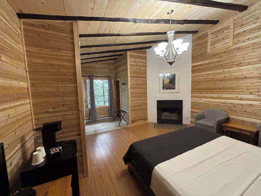Een houten hutje van binnen met een bed, een open haard, een kroonluchter en uitzicht door een raam.