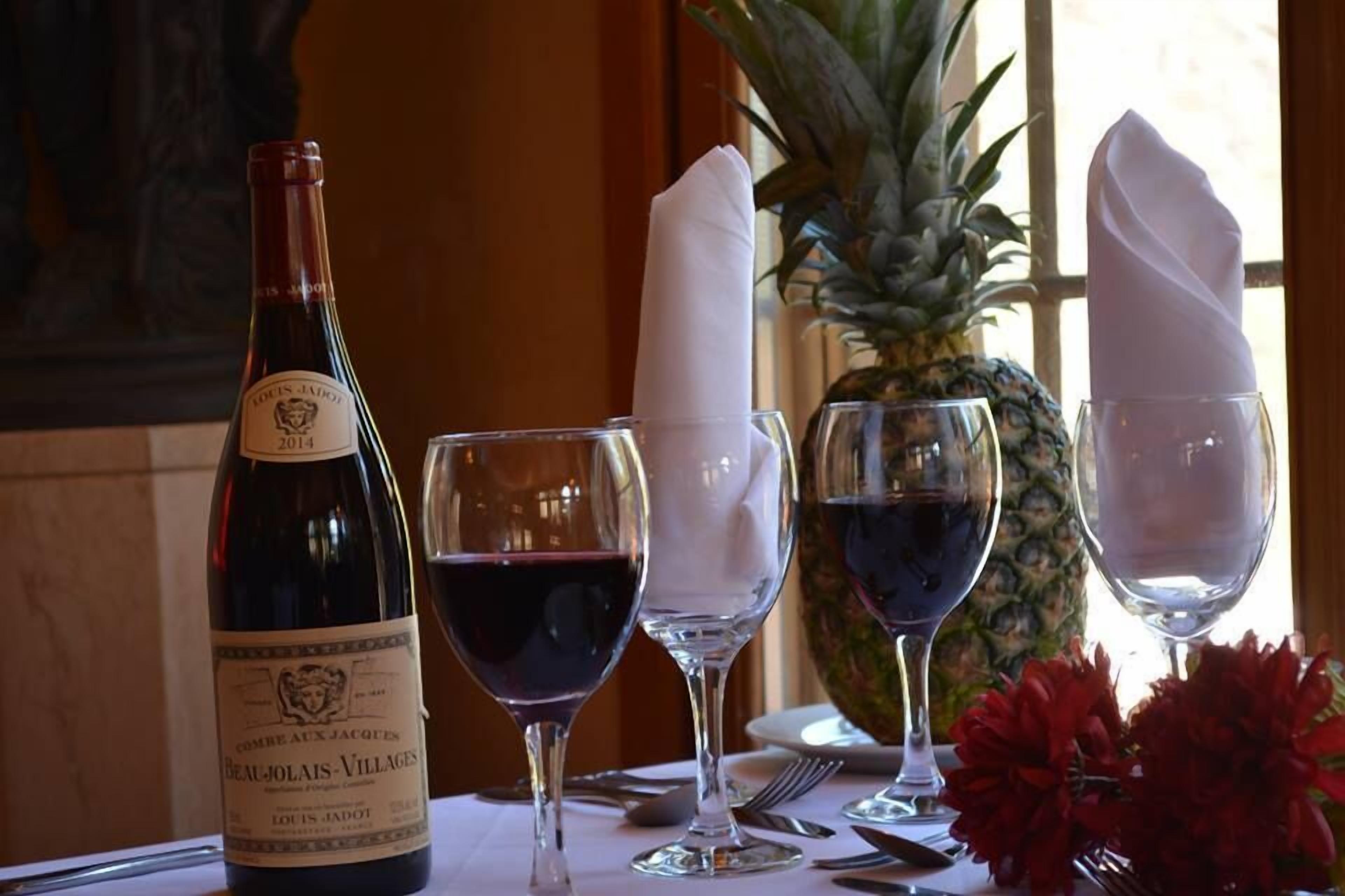 Eine Flasche Louis Jadot Wein, zwei Rotweingläser und eine Ananas auf einem Tisch mit weißen Servietten und roten Blumen.