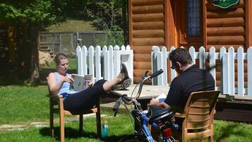 Two people sitting at an outdoor table with a bicycle, in front of a wooden cabin with a white picket fence.