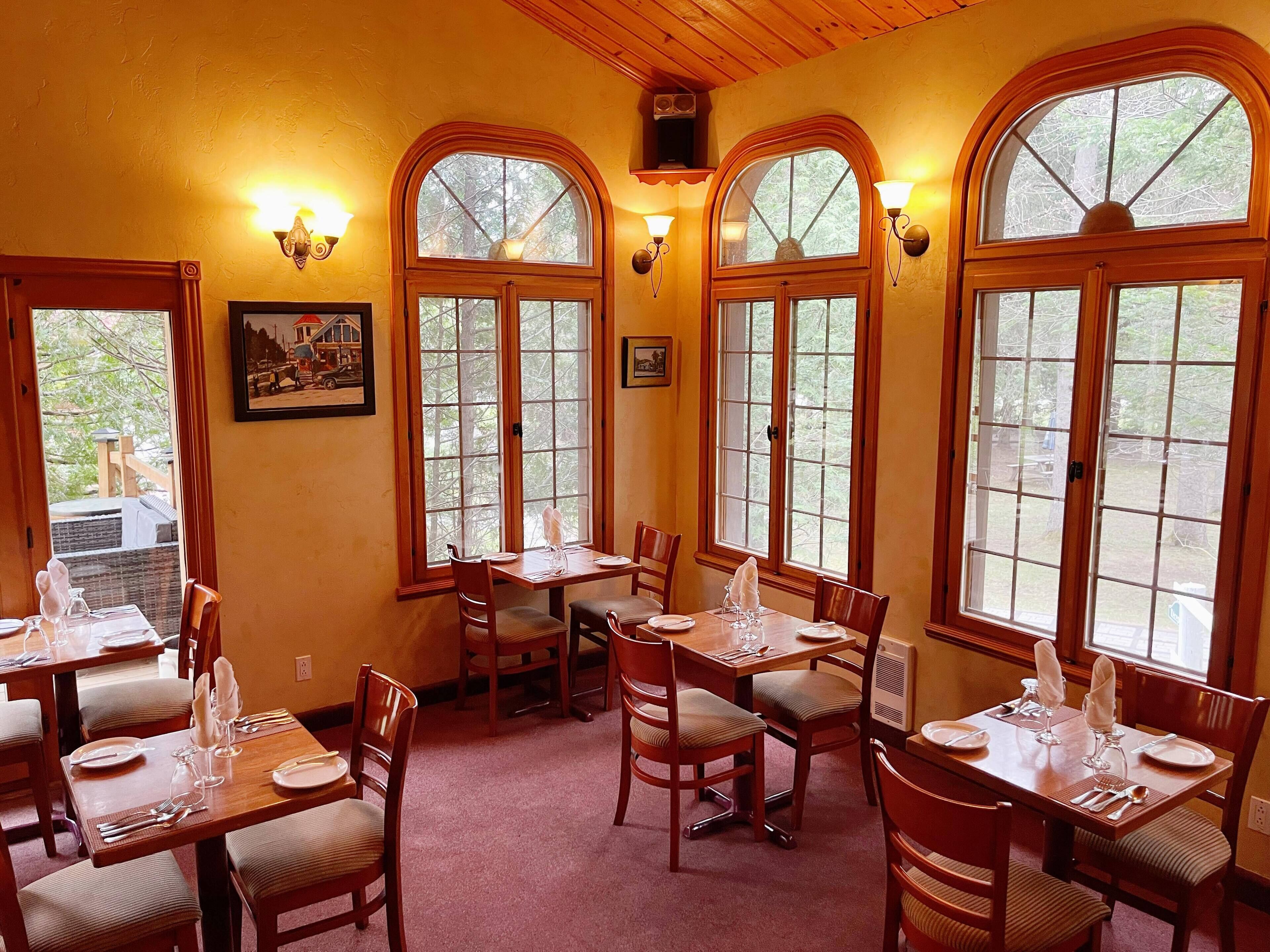 A dining area with wooden tables and chairs, large arched windows, and wall-mounted light fixtures.