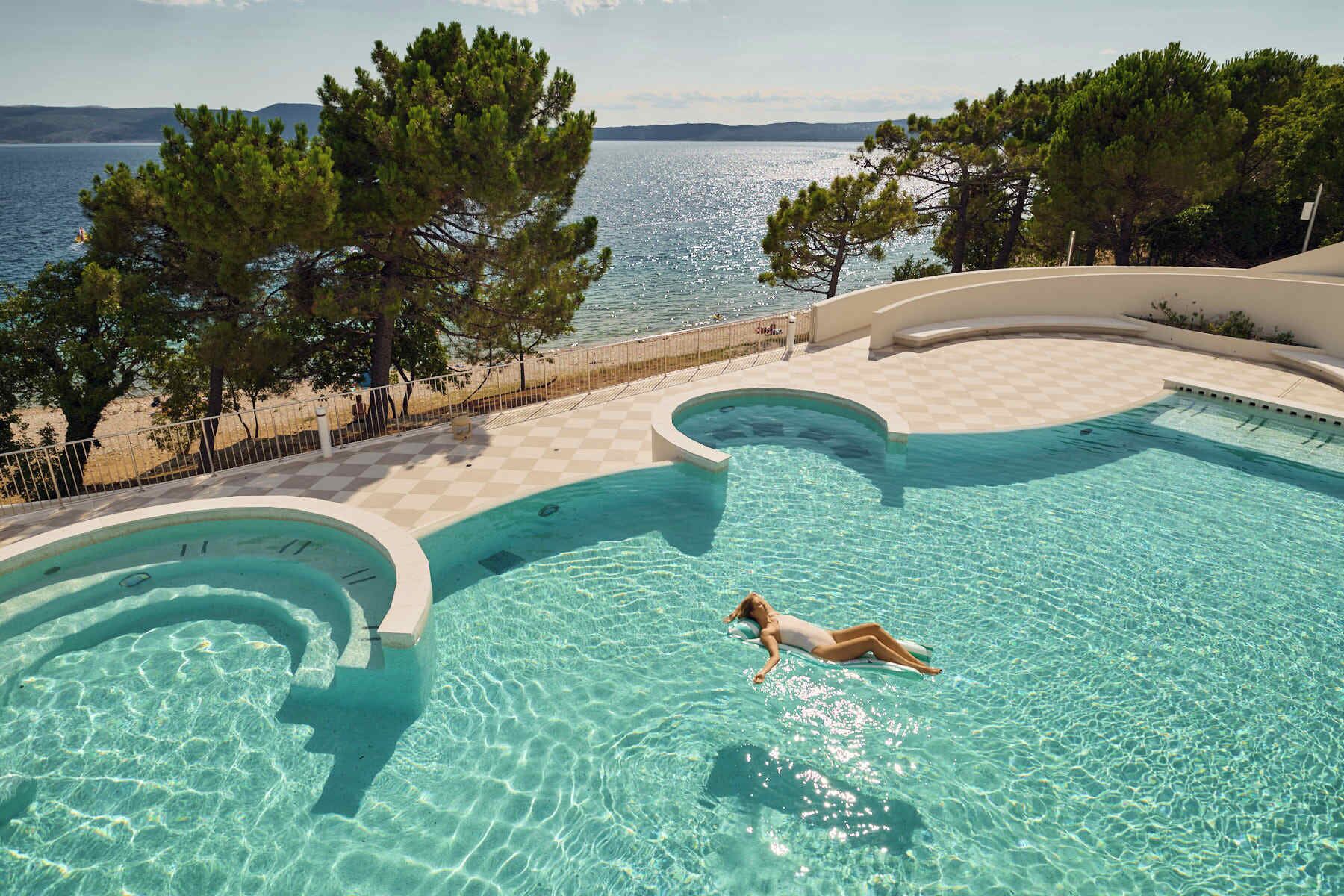 Una piscina con una persona flotando, rodeada de árboles y vista al mar.